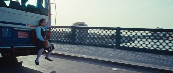 Film still of a little girl sitting on the back of a bus, going over a bridge.