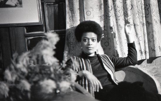 Black and white photo of a Black woman with a short afro sitting in a chair in front of curtains.