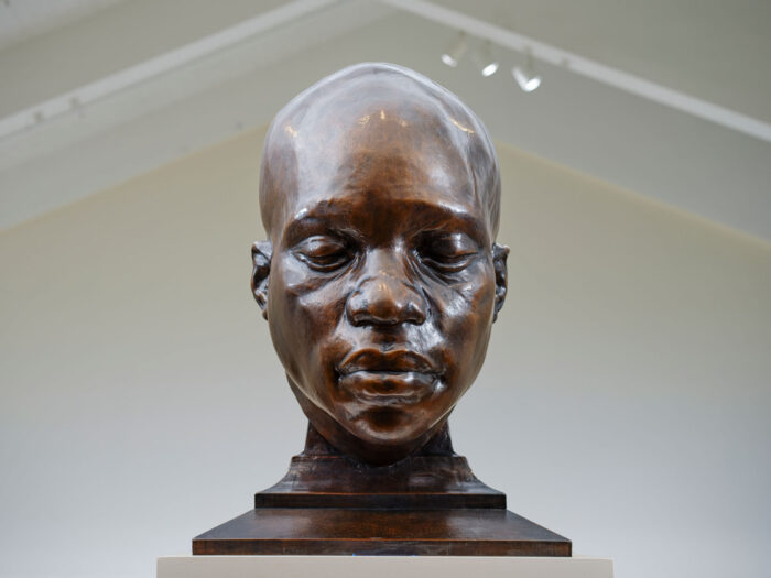 A bronze sculpture of a Black man's face and neck. His eyes are closed.