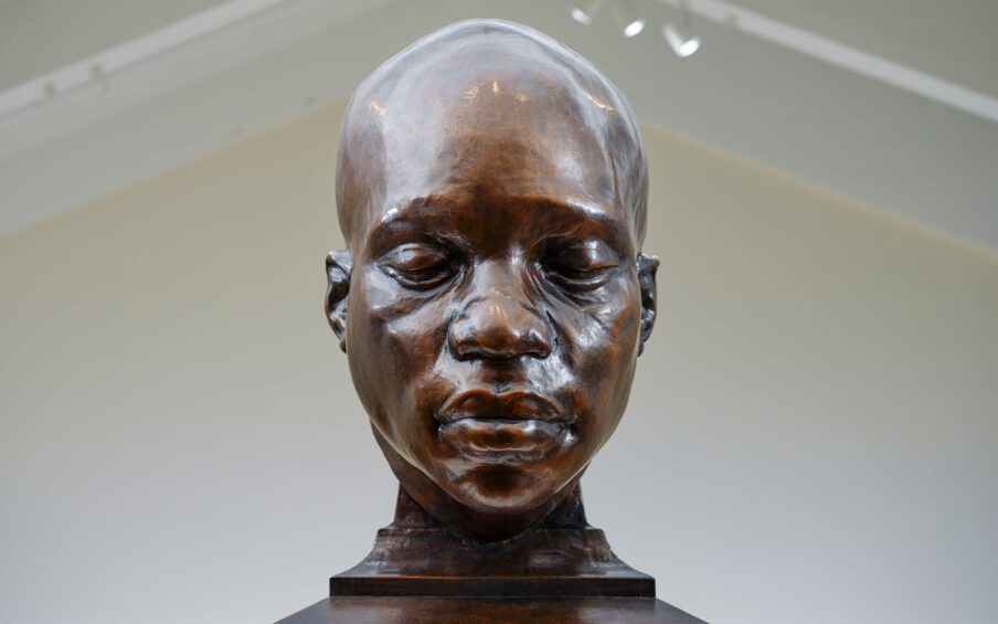 A bronze sculpture of a Black man's face and neck. His eyes are closed.