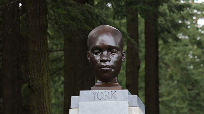 A bronze-looking head statue of a Black man with his eyes closed. It's on a stone pedestal and surrounded by big trees.
