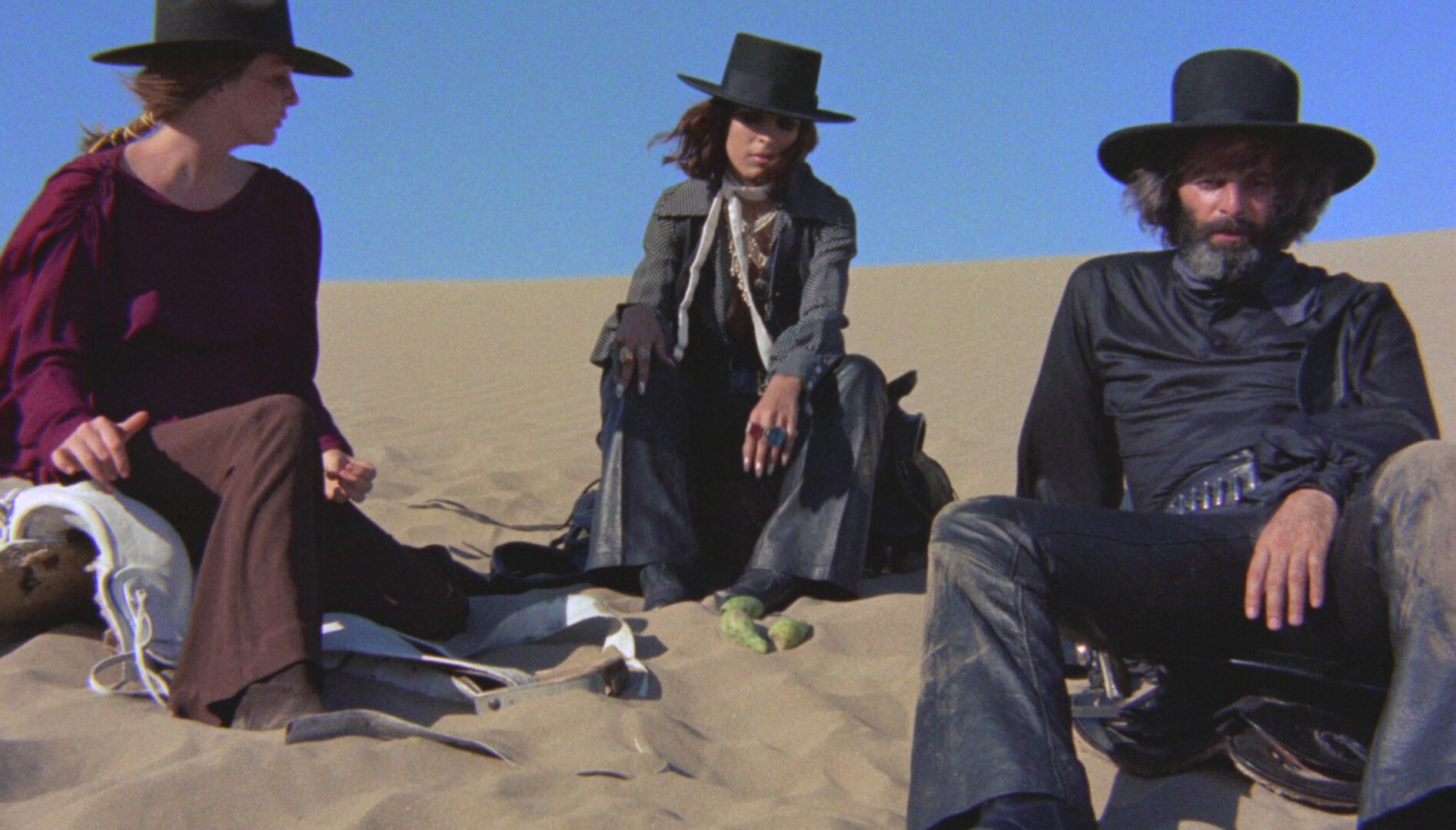 Film still of three people in wide-brimmed hats, sitting on sand.