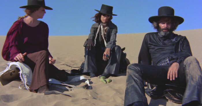 Film still of three people in wide-brimmed hats, sitting on sand.