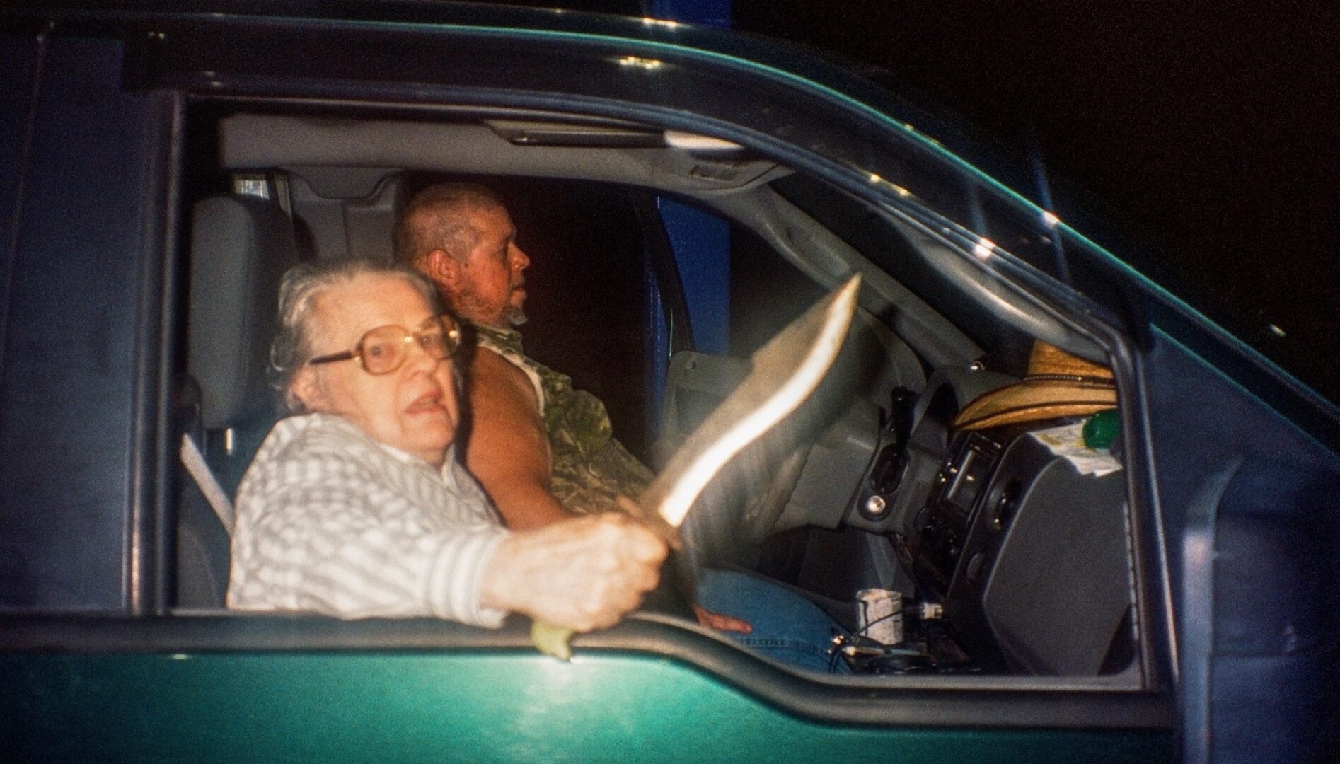 FIlm still of a woman holding a big knife sitting in the passenger seat of a green truck.