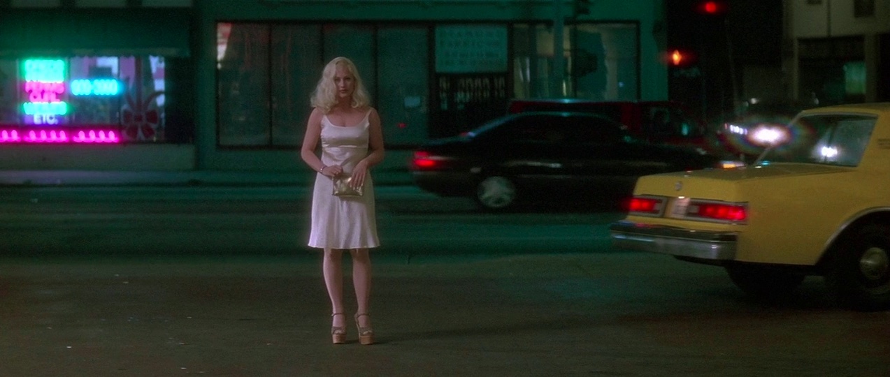 Film still of a woman with blonde hair and a pink dress, standing on a city street at night.