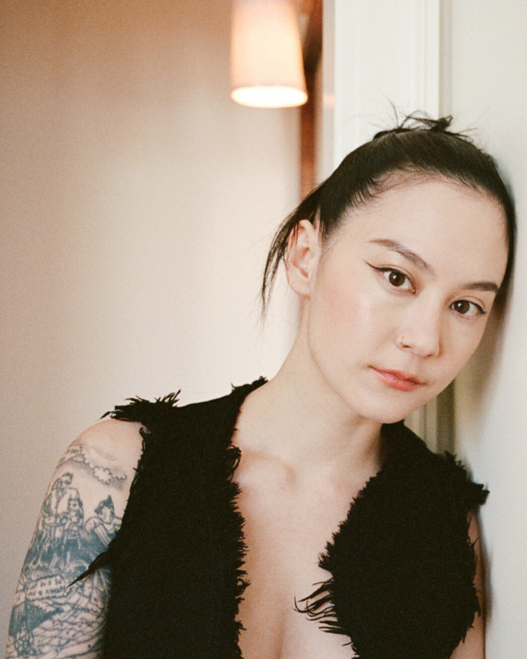 Photo of a woman wearing a black vest, with tattoos on her arm, leaning against a wall.