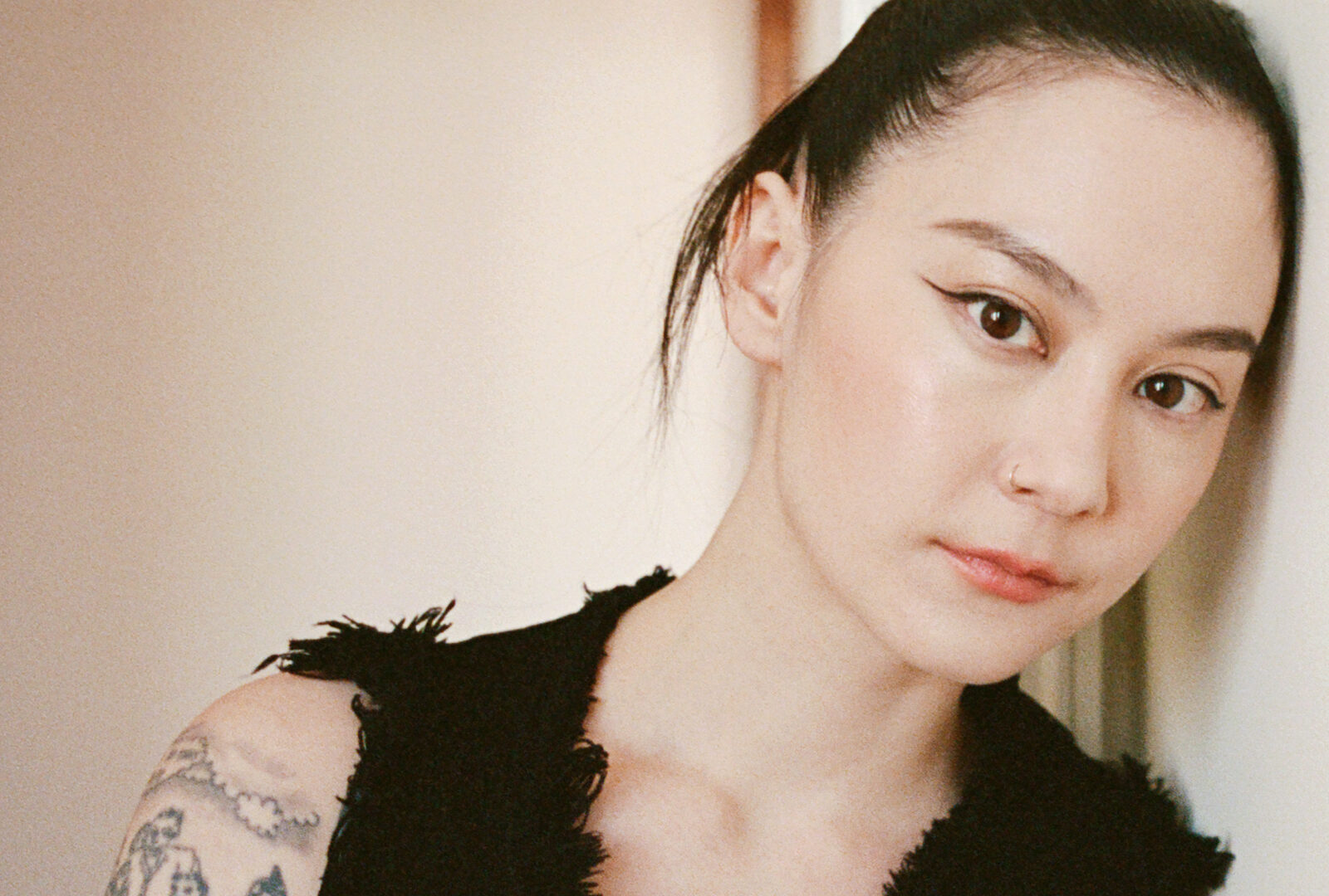 Photo of a woman wearing a black vest, with tattoos on her arm, leaning against a wall.