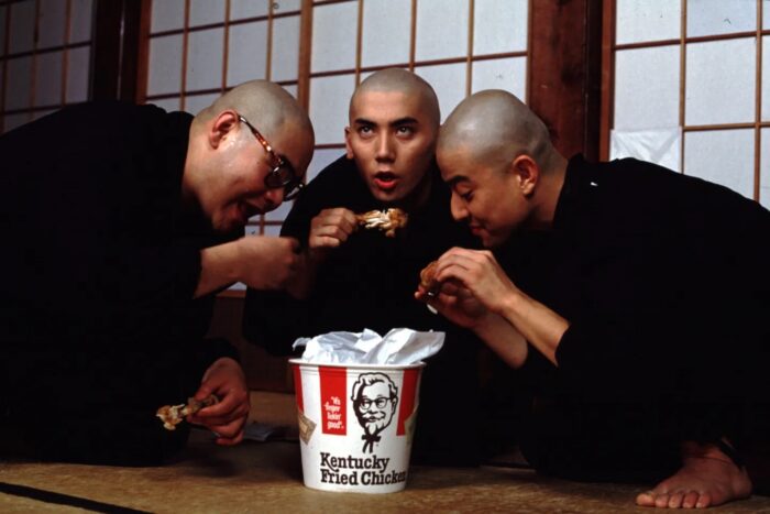 Three men with shaved heads and monk's robes kneeling and eating Kentucky Fried Chicken.