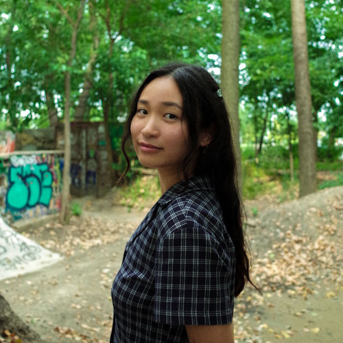 Portrait of a woman with long brown hair standing in front of trees and a graffitied wall.
