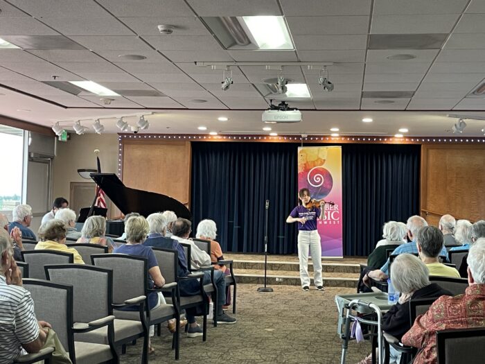Photo of a young violin player playing to a room of seated people.