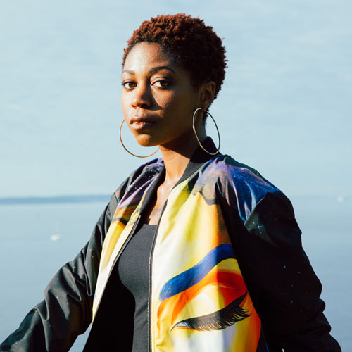 Portrait of a Black woman with short hair, wearing large hood earrings and a space-themed jacket, standing in front of a body of water.