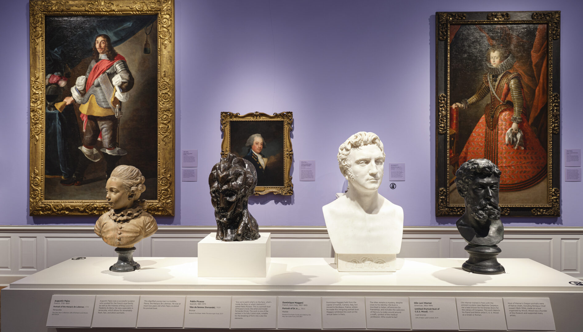 Photo of a gallery with purple walls, paintings on the walls and four bust sculptures in the foreground.