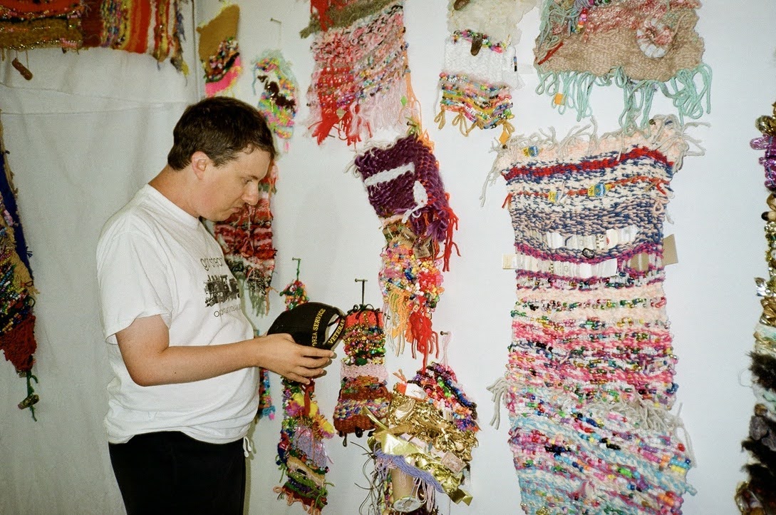Photo of a man in a white t-shirt, holding a black baseball hat, standing in front of a wall hung with colorful weavings.