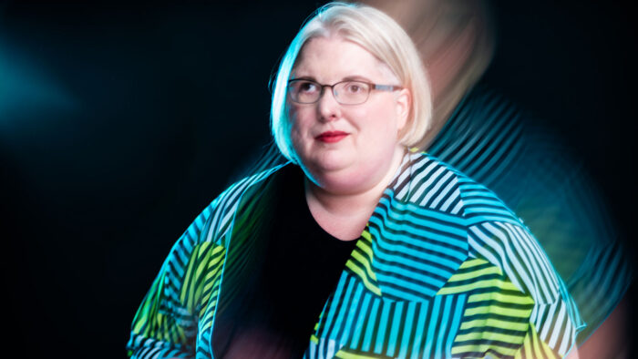 Photo of a person with blonde hair and eyeglasses wearing a striped jacket over a black shirt. They are against a black background.