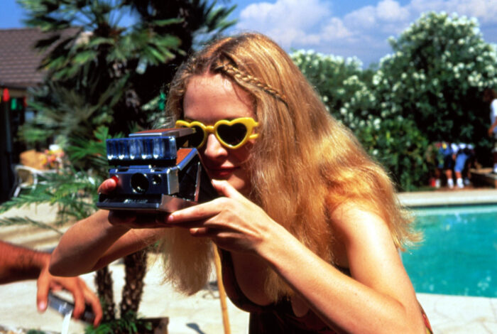 A woman with long blonde hair, wearing heart-shaped sunglasses and holding a camera up to her face.