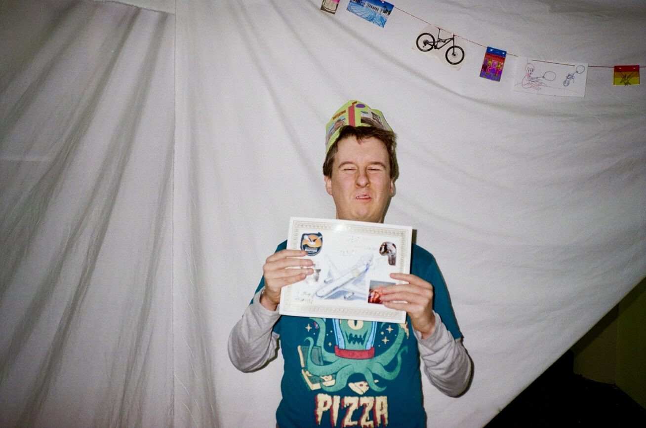Photo of a smiling man wearing a paper crown and holding up a certifcate. He is wearing a t-shirt with an alien on it that says Pizza.