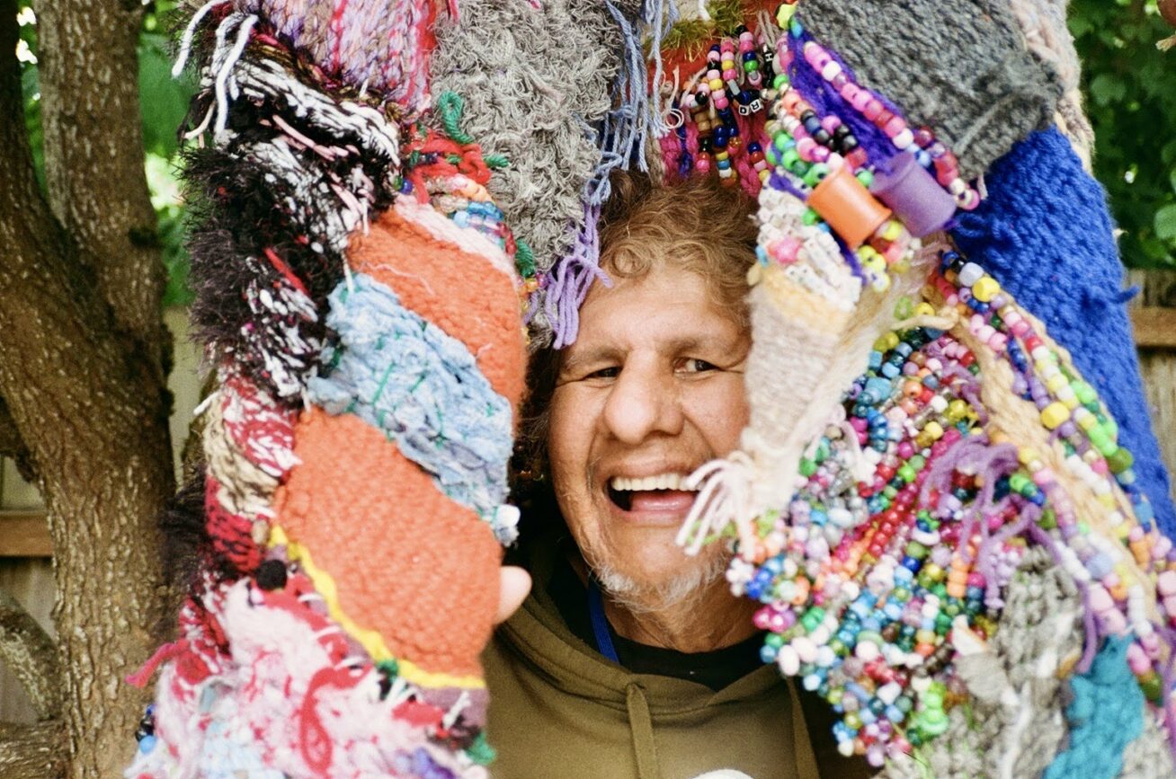 Photo of a smiling man behind some colorful fabric weavings