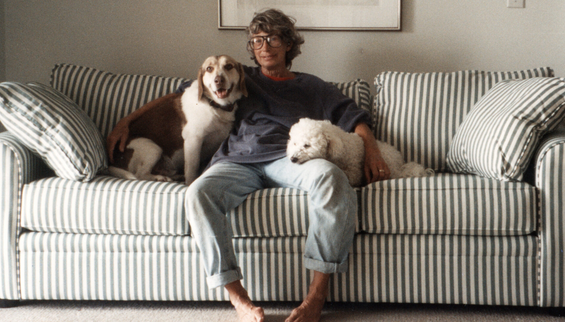 Image of Mary Oliver sitting barefoot on a striped sofa with dogs on either side of her.