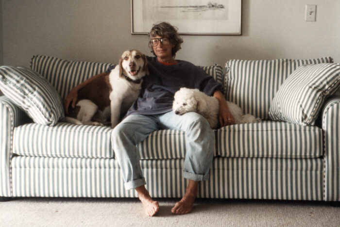 Image of Mary Oliver sitting barefoot on a striped sofa with dogs on either side of her.
