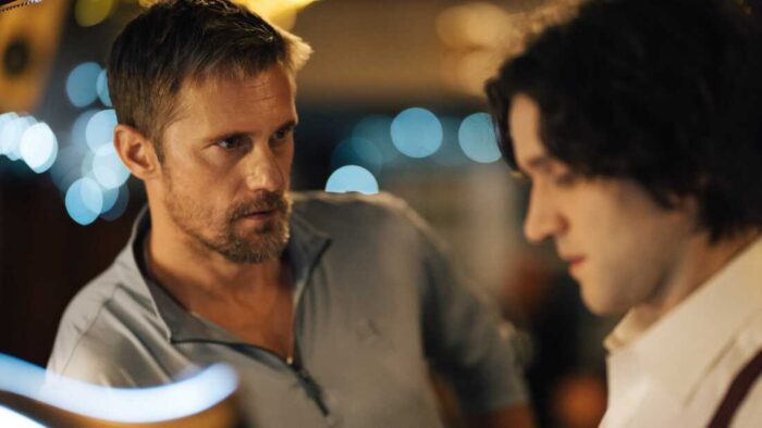 Film still of a blond man with facial hair staring intently at a young man with curly brown hair.