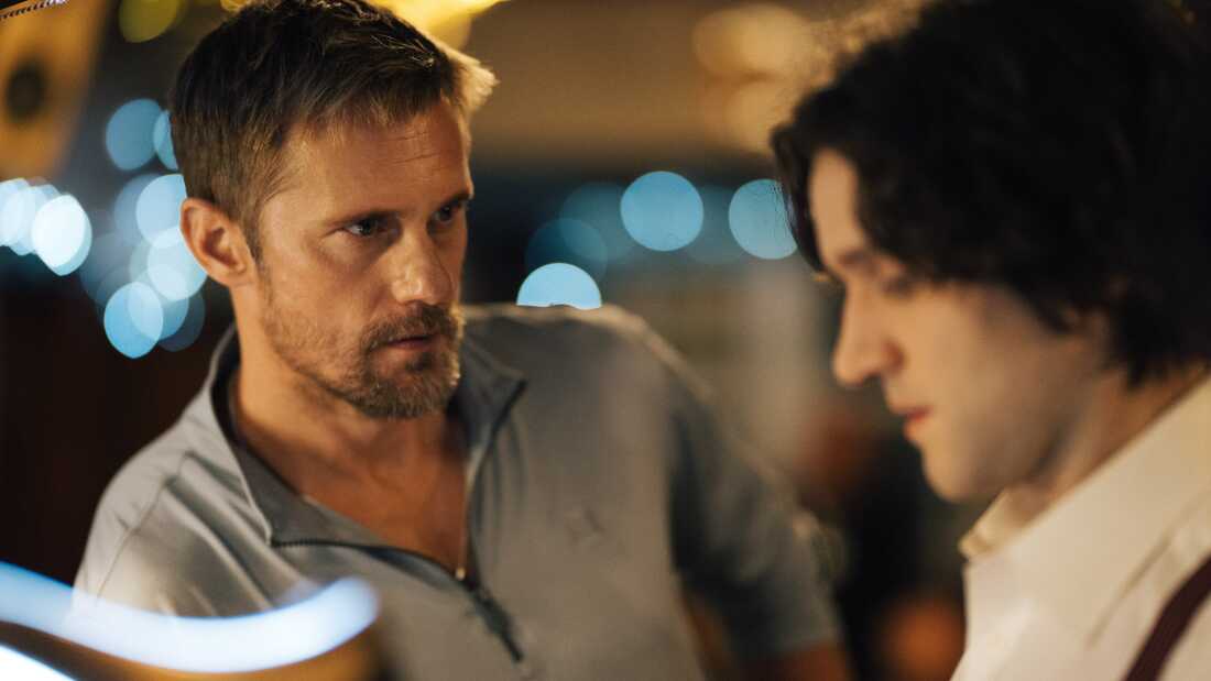 Film still of a blond man with facial hair staring intently at a young man with curly brown hair.