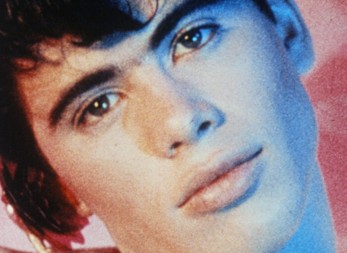 Close-up film still of a young man's face. He has brown hair and his head tilted to the side.