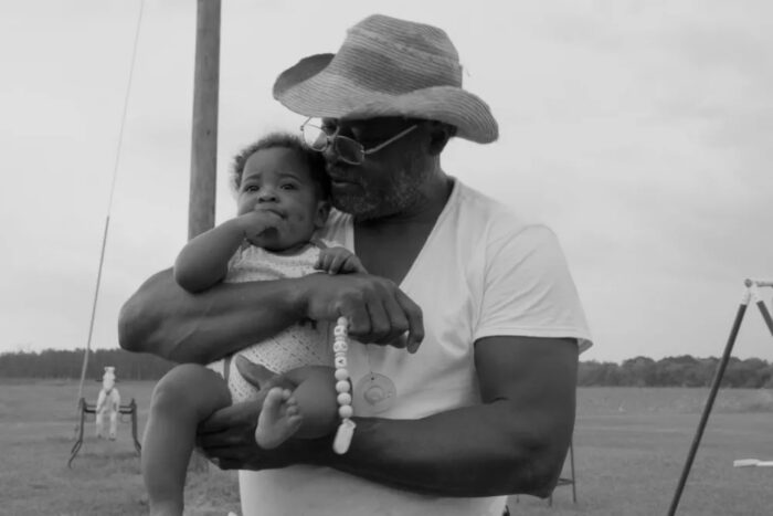 A black and white photo of a black man in a cowboy hat, holding a baby and kissing its cheek.