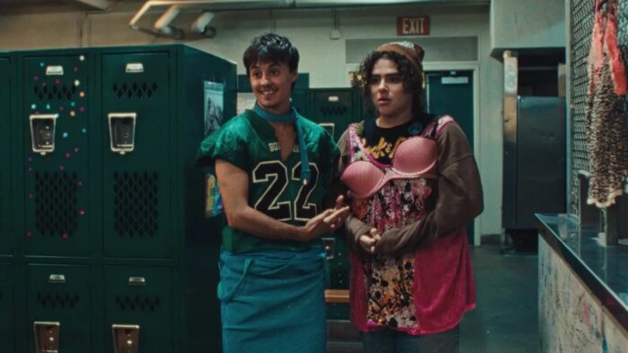 A photo of two teenagers in a locker room. One has a football jersey on and the other is wearing a pink bra outside of their shirt.