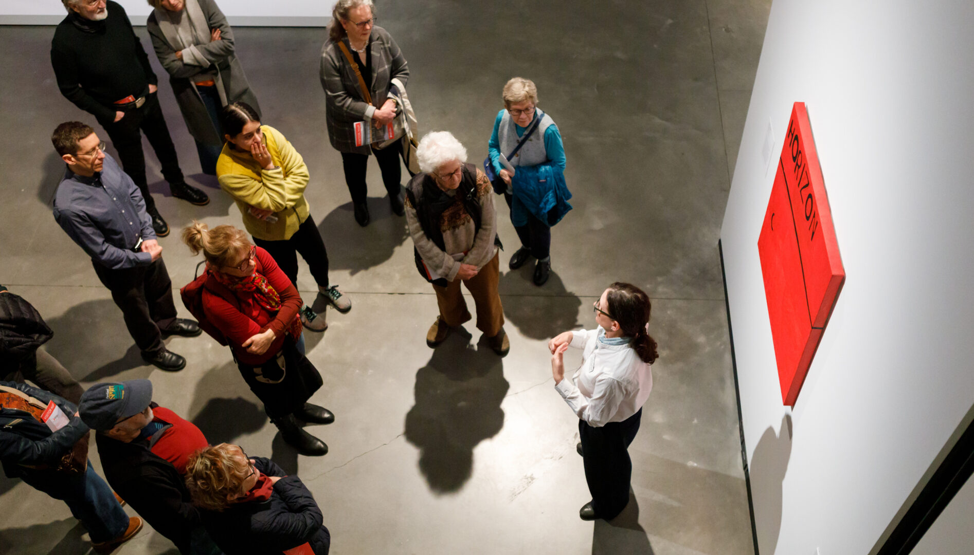 A group of people standing around a painting. A curator is in front of the painting speaking to the crowd.