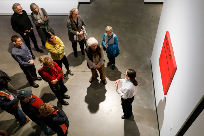 A group of people standing around a painting. A curator is in front of the painting speaking to the crowd.