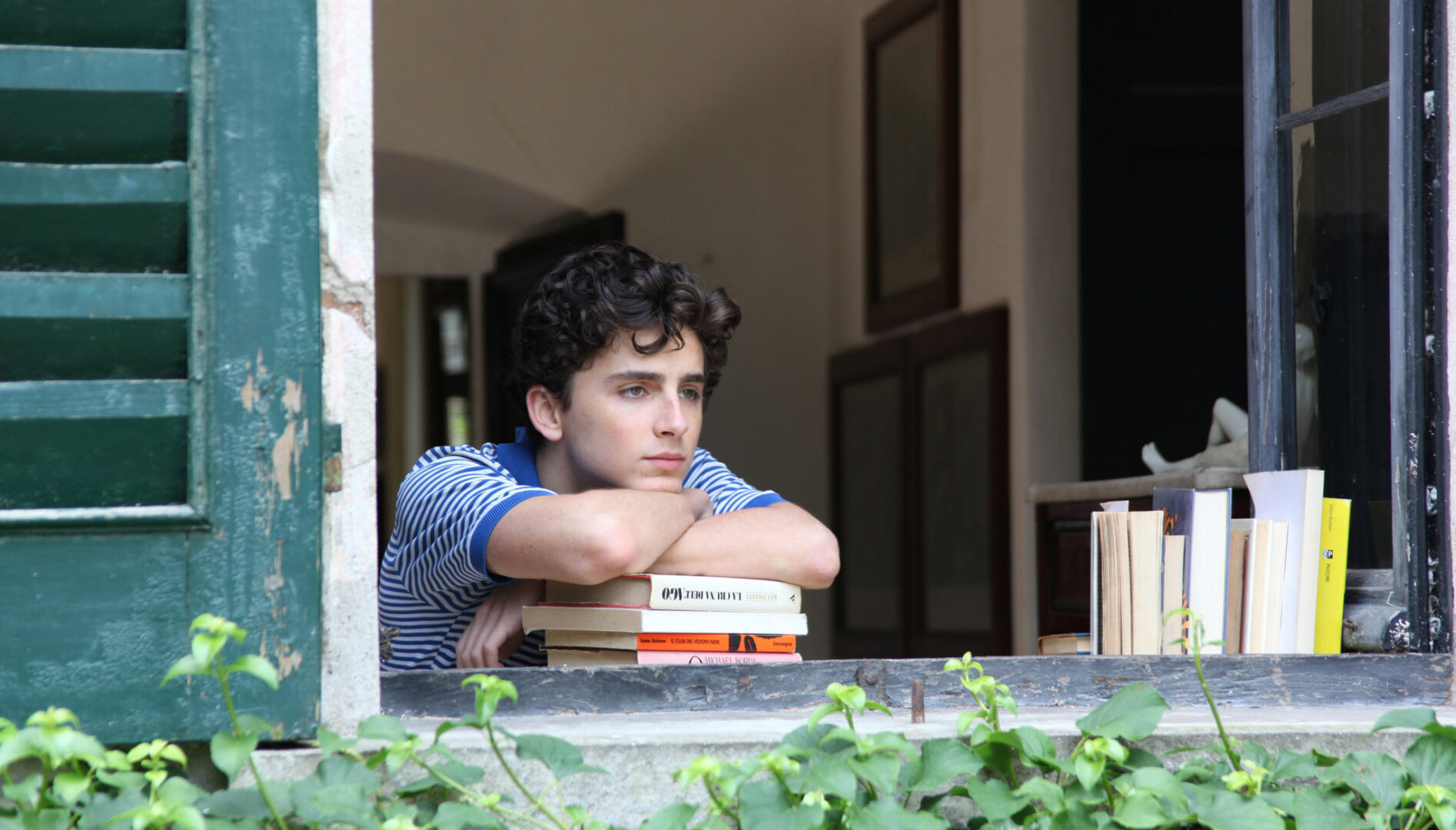 Film still of a young man looking out a window with his head and arms resting on a pile of books.
