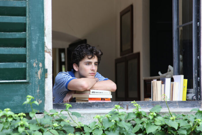 Film still of a young man looking out a window with his head and arms resting on a pile of books.