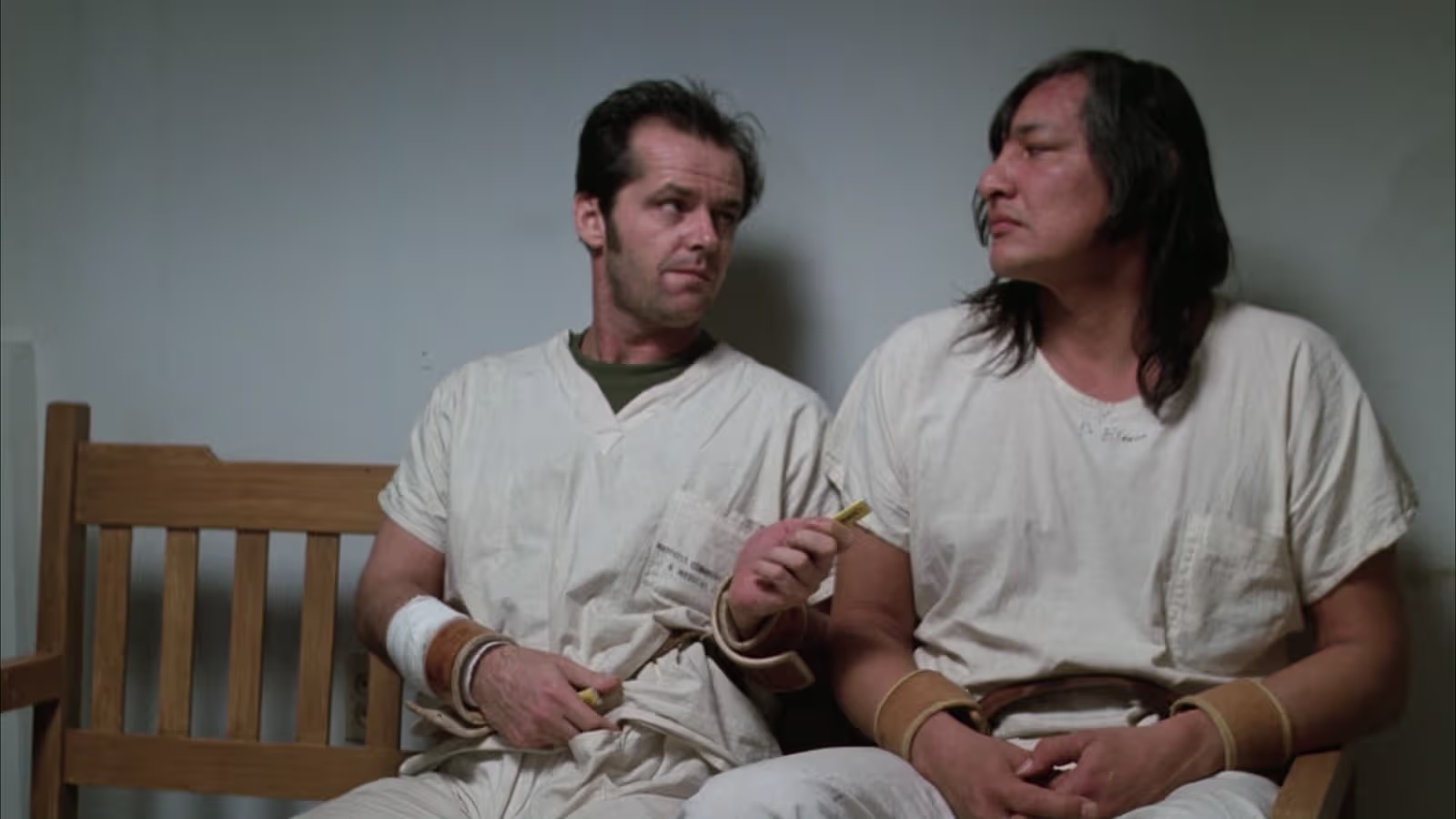 Two men sitting on a bench wearing white hospital clothes. They are looking at each other and one is offering a piece of gum to the other.