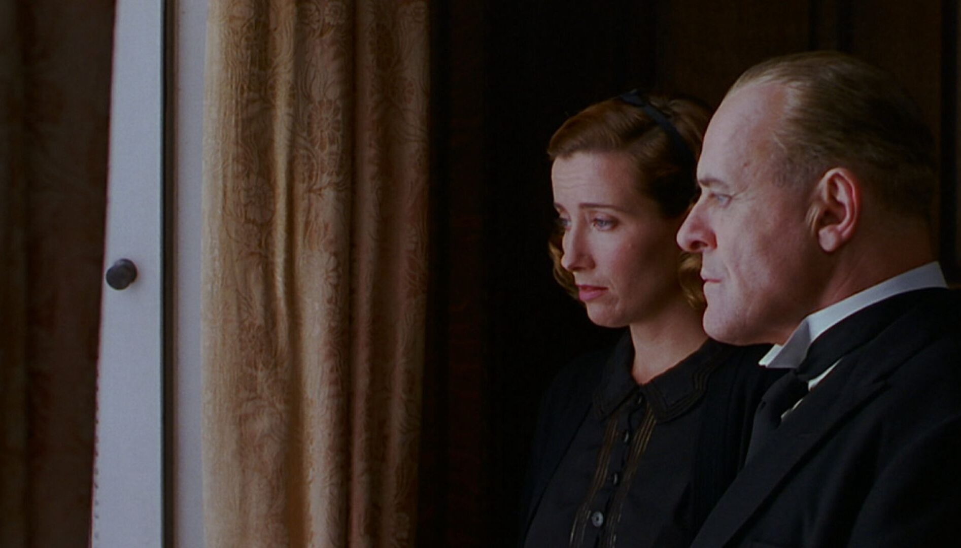 A man and a woman with serious faces looking out a window.