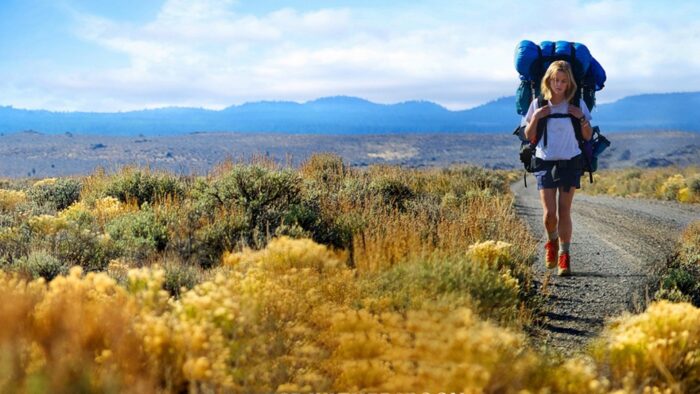A young woman carrying a large backpack, hiking through a beautiful landscape with mountains in the background.