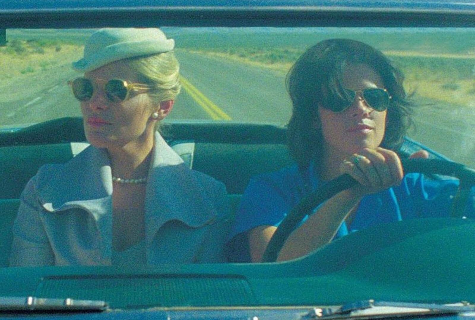 Two women wearing sunglasses, driving in a convertible on a desert road.