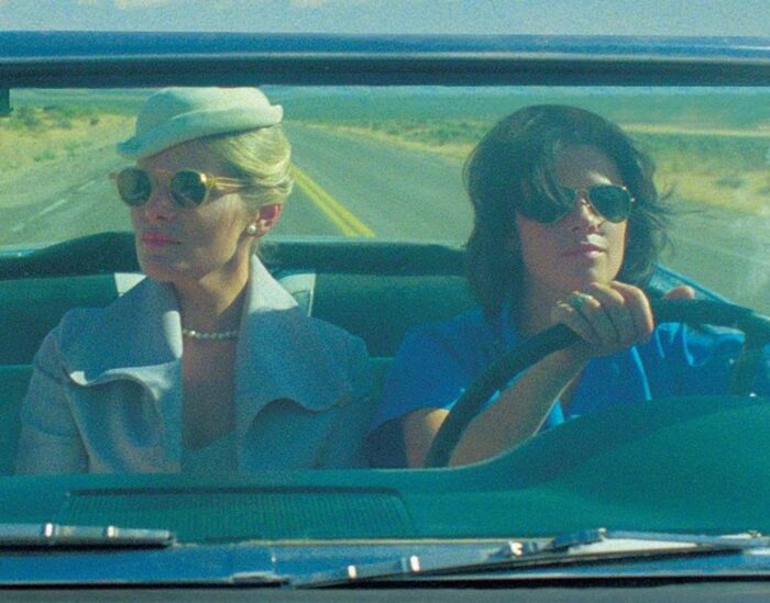 Two women wearing sunglasses, driving in a convertible on a desert road.