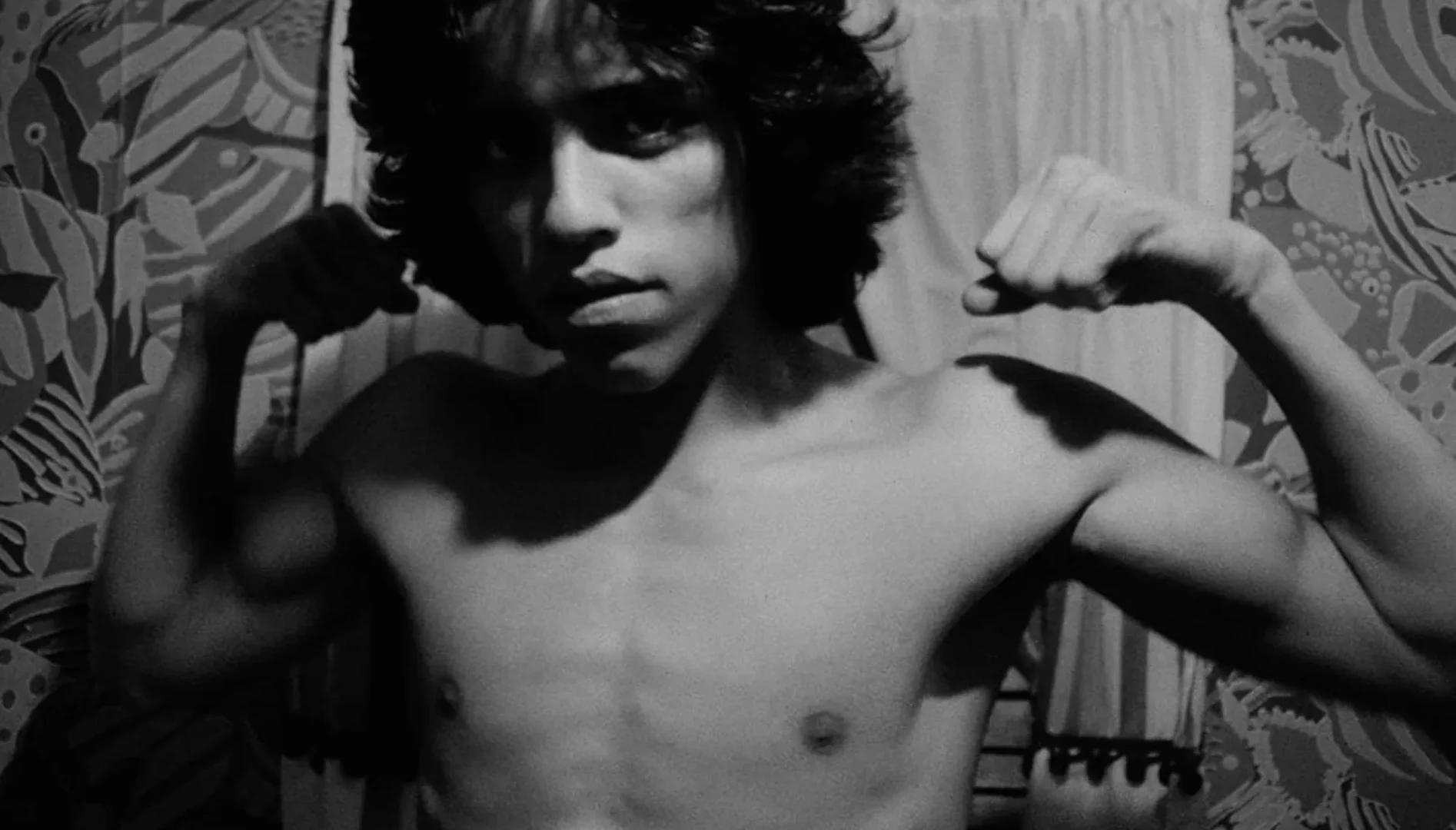 Black and white film still of a young, shirtless man holding up his arms in a muscle pose.