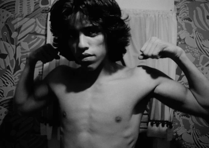 Black and white film still of a young, shirtless man holding up his arms in a muscle pose.
