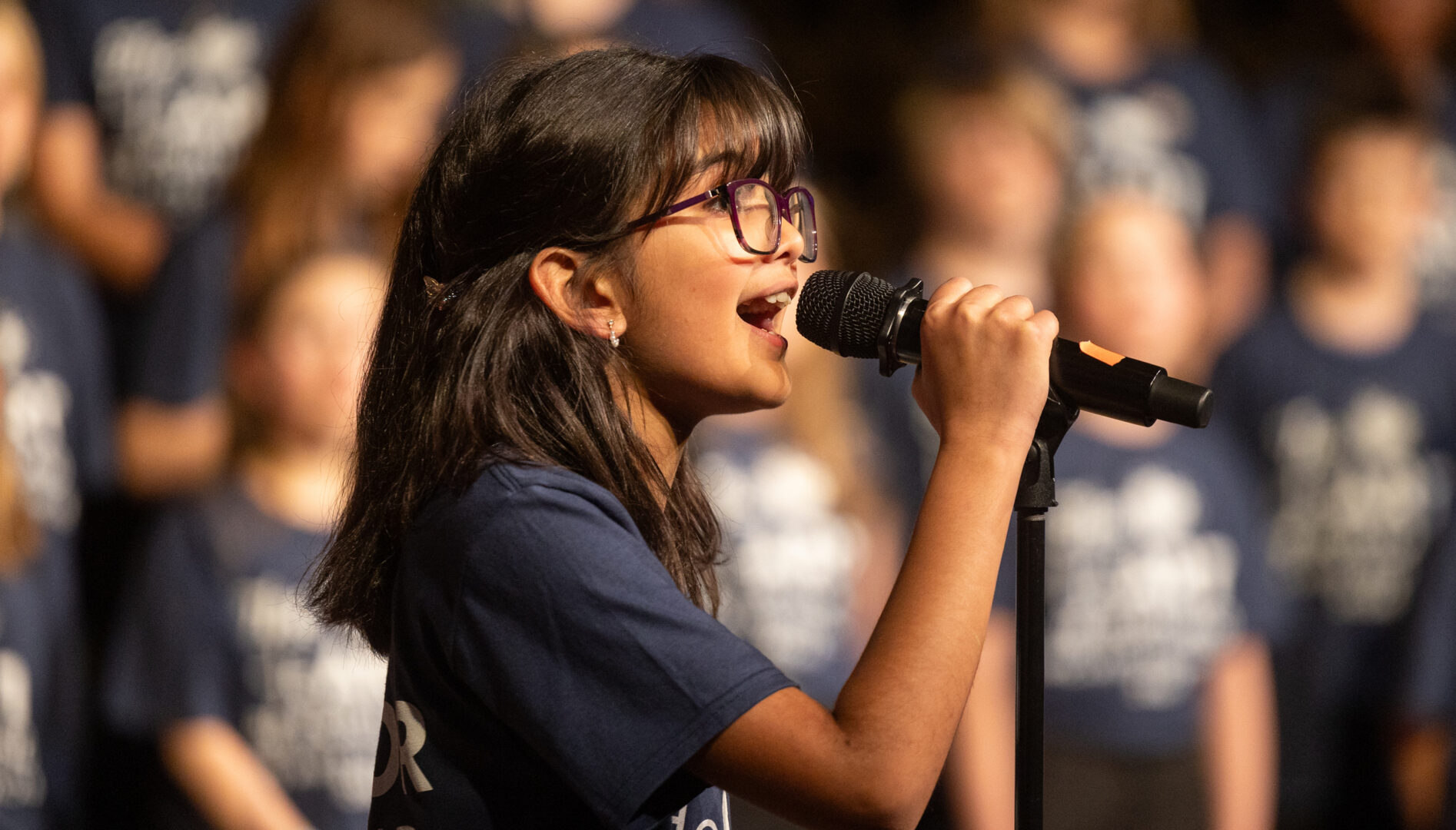 A young girl in a dark blue tee shirt singing into a microphone.