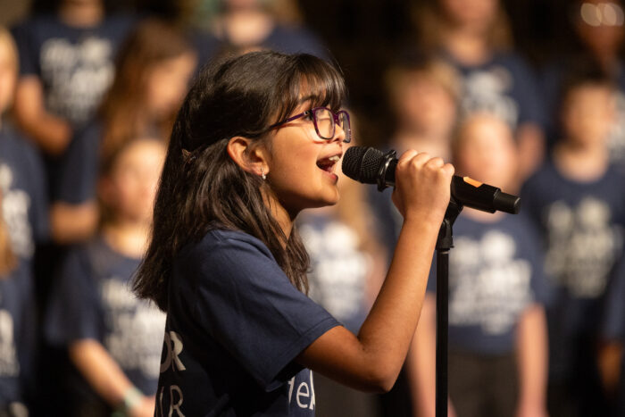 A young girl in a dark blue tee shirt singing into a microphone.