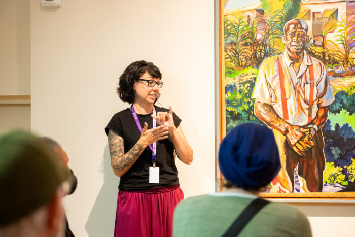 Woman doing sign language in front of a painting by Isaka Shamsud Din.