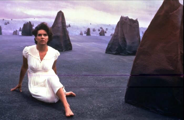 A woman in a white dress sitting on a beach surrounded by rocks.
