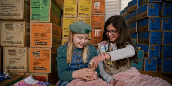 Two young girls, wearing Girl Scout uniforms, sitting in front of stacks of cookie boxes.