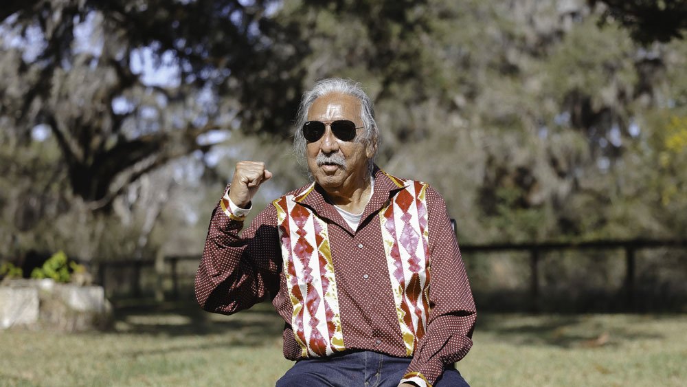 Leonard Peltier, wearing sunglasses and a maroon, patterned shirt, sitting outside in front of trees.