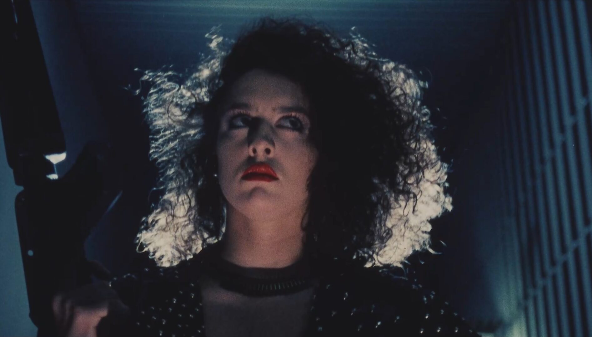 Film still of a backlit woman with big curly hair, holding a large gun.
