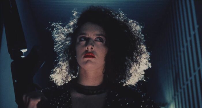Film still of a backlit woman with big curly hair, holding a large gun.