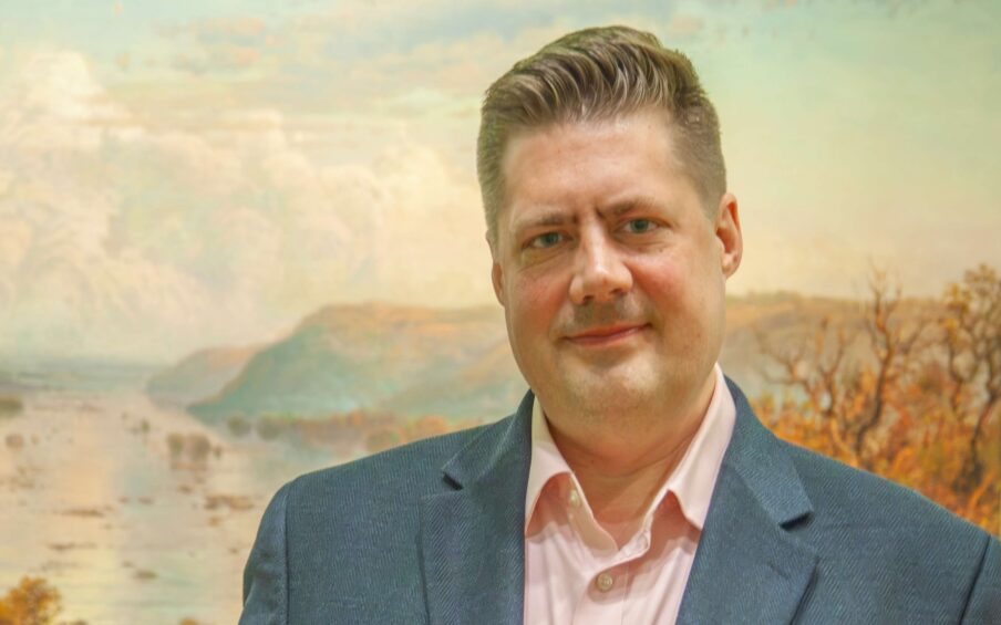 Photo of a man wearing a blue suit jacket and a pink Oxford shirt, standing in front of a landscape painting.