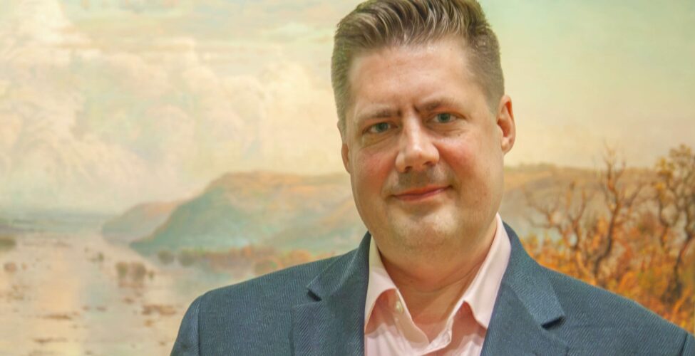 Photo of a man wearing a blue suit jacket and a pink Oxford shirt, standing in front of a landscape painting.