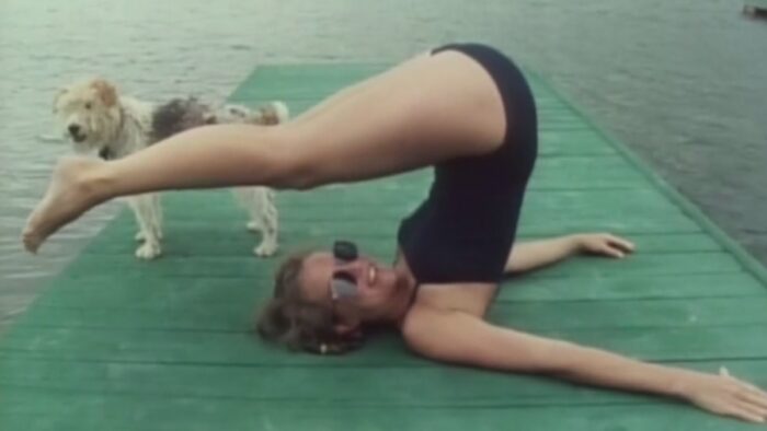 Film still of a woman on a boat dock doing an acrobatic pose. She is wearing a black bathing suit and sunglasses. A dog is standing behind her.